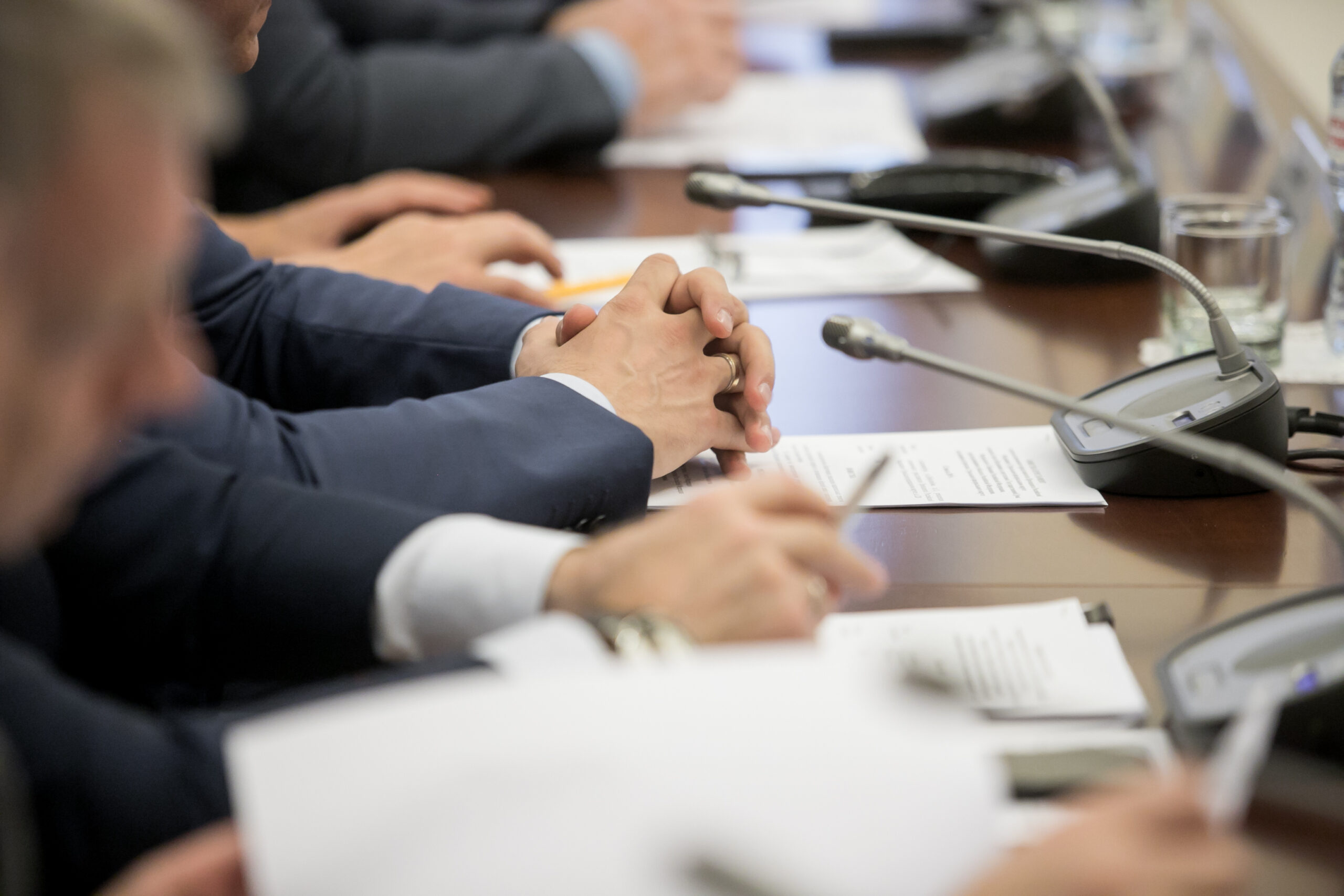 Das Bild zeigt eine Szene aus einer geschäftlichen Konferenz, bei der Männer in schicken Anzügen an einem Rednerpult sitzen. Die Hände der Männer sind im Fokus, was auf eine aktive Beteiligung an Diskussionen, Präsentationen oder Besprechungen hinweist.